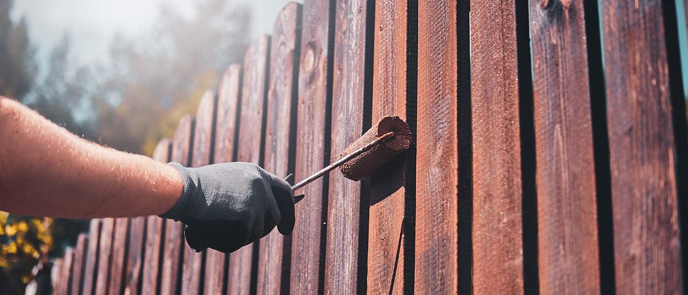 A person painting the side of a wooden fence.